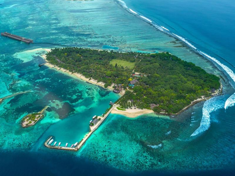 Kleine Insel mit dichtem Grün, umgeben von türkisblauem Wasser und Korallenriffen, mit Steg und Bootshäusern.