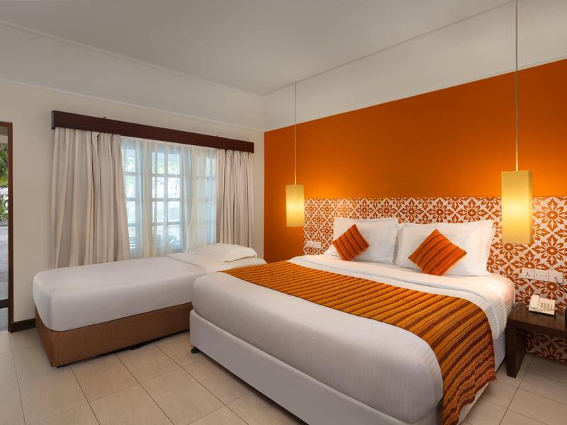 Hotelzimmer mit großem Bett, Einzelbett, oranger Wand und zwei Hängelampen, heller Boden und Vorhänge.