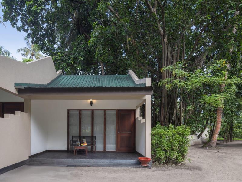 Modernes Haus mit weißer Fassade, grünem Dach und Holztür, umgeben von Bäumen und Pflanzen.