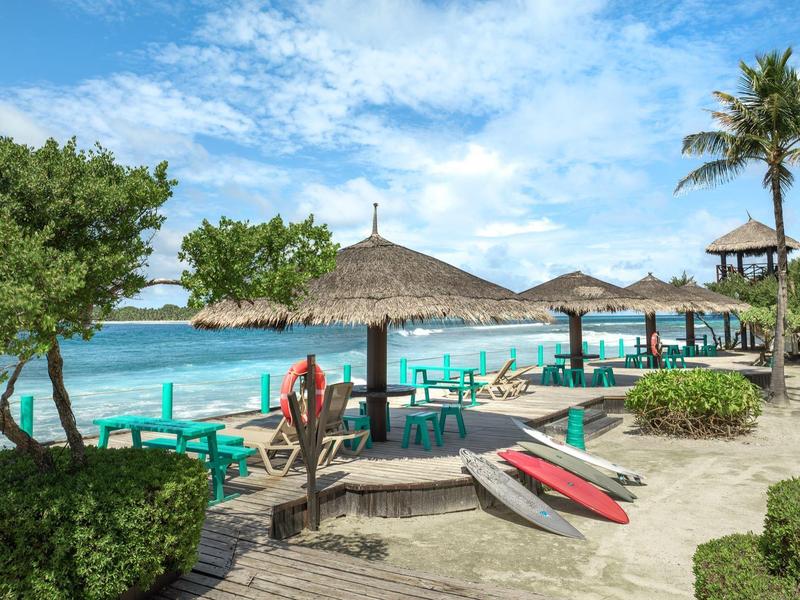 Strand mit türkisfarbenem Wasser, Palmen, Strohdächern und bunten Liegestühlen am Holzsteg.
