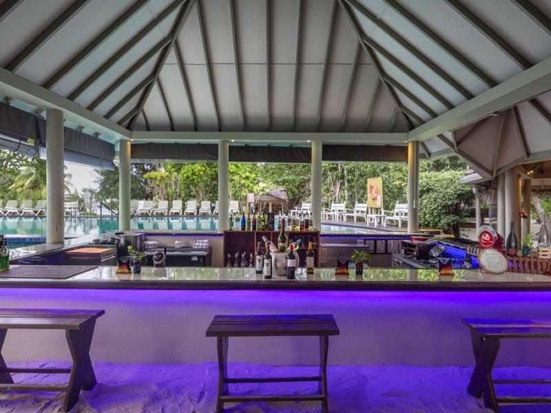 Offene Bar mit Holzhockern, blauer LED-Beleuchtung und Blick auf Wasser und Palmen im Hintergrund.