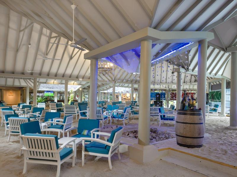 Helle Strandbar mit weißem Holz, türkisfarbenen Stühlen und Sandboden unter einem offenen Dach.