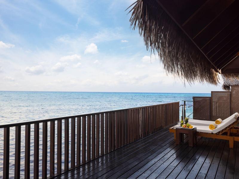 Holzterrasse mit Liegestuhl und Tisch am Meer unter blauem Himmel mit Wolken.