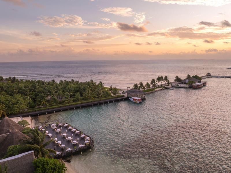 Inselresort mit Holzdocken, Palmen und Liegestühlen am klaren Meer bei Sonnenuntergang.