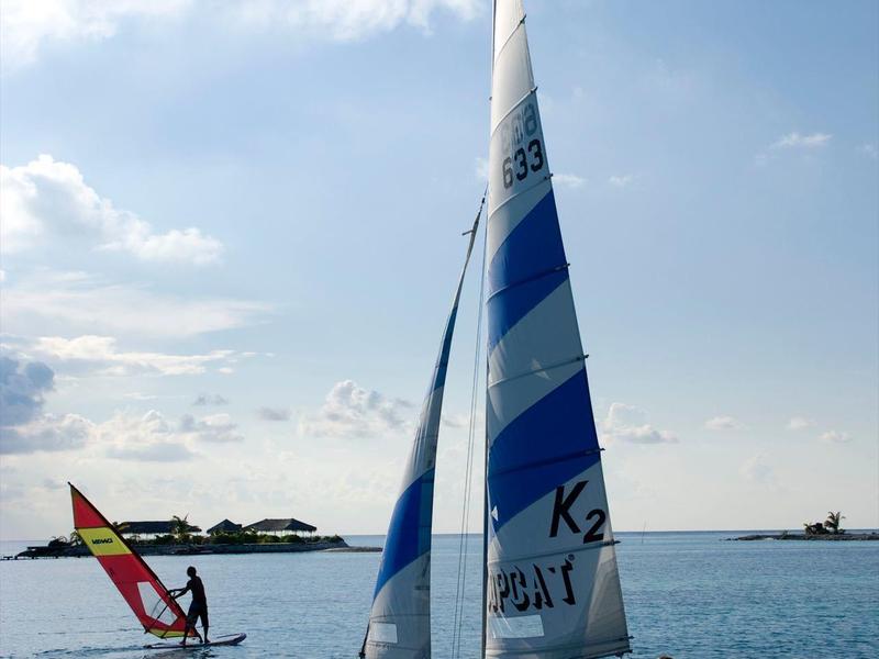 Segelboot mit blau-weißen Segeln auf ruhigem Wasser, Hintergrund mit Wolken und Windsurfer.