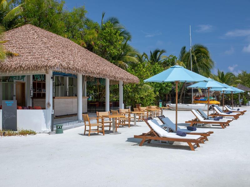 Strand mit weißen Liegestühlen, blauen Sonnenschirmen und einem Strohdach-Pavillon vor Palmen.