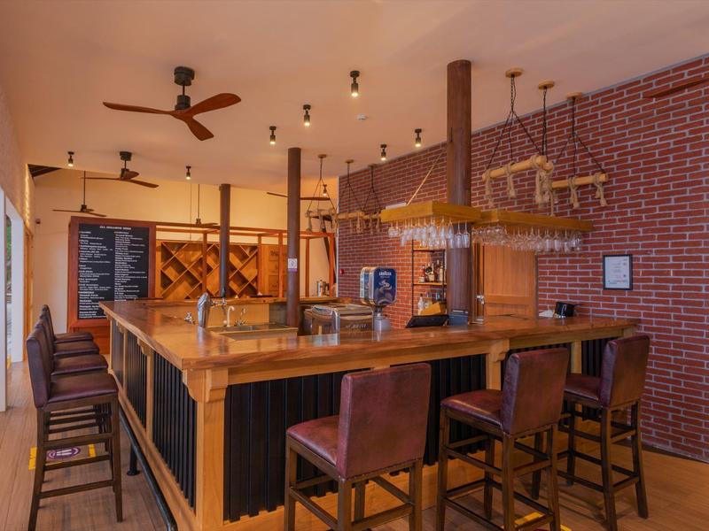 Innenraum einer Bar mit Holztresen, hohen Stühlen, Deckenventilator und roter Ziegelwand.