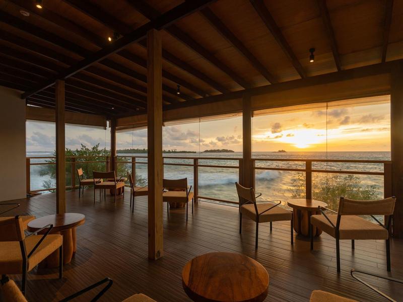 Offene Terrasse mit Holzboden und Möbeln, Blick auf Meer und Sonnenuntergang am Horizont.