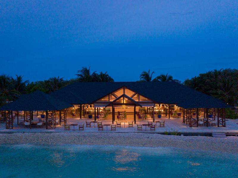 Ein beleuchtetes Strandrestaurant mit offenem Holzgestell bei Dämmerung am Ufer.