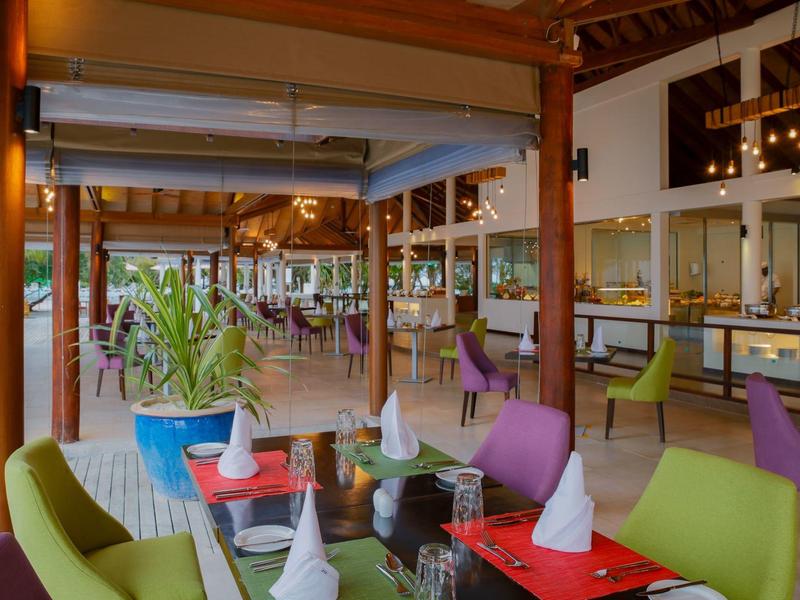 Modernes, offenes Restaurant mit bunten Stühlen, Holzbalken, grünen Pflanzen und gedeckten Tischen.