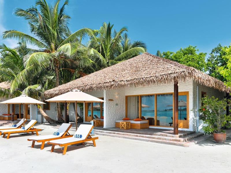 Strandbungalow mit Strohdach, Liegestühlen und Palmen unter klarem blauem Himmel.