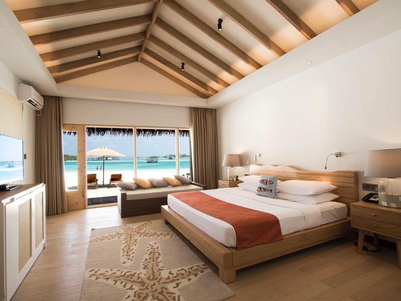 Helles Hotelzimmer mit großem Bett, Holzdecke, TV und Balkon mit Meerblick und Liegestühlen.