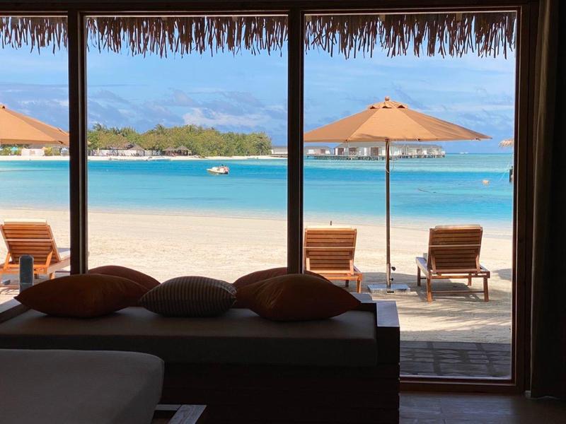 Blick aus einem Zimmer auf Strand mit Sandalen, Sonnenschirm und blauem Meer unter wolkenlosem Himmel.