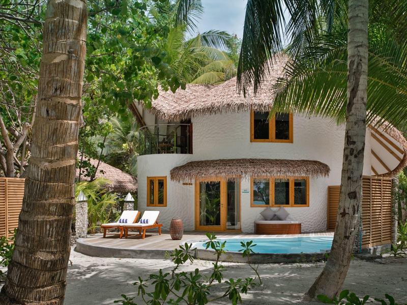Weißes Haus mit Strohdach, umgeben von Palmen und grünem Laub, mit Terrasse und Pool.