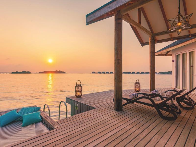 Sonniger Holzsteg mit Liegestühlen, Kissen und Laternen, Blick auf ruhiges Meer bei Sonnenuntergang.