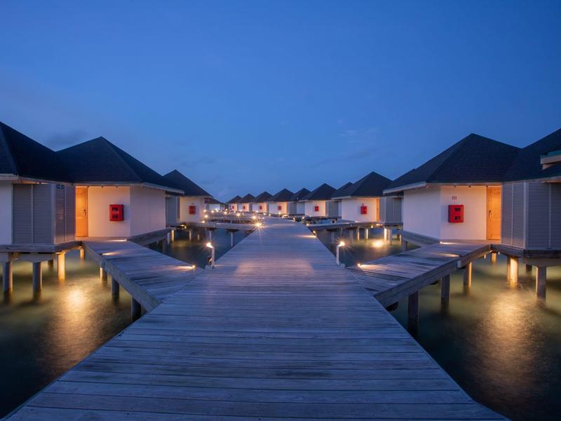 Holzhäuser auf Stelzen mit breitem Steg am Wasser bei Blauhour, helle Beleuchtung.