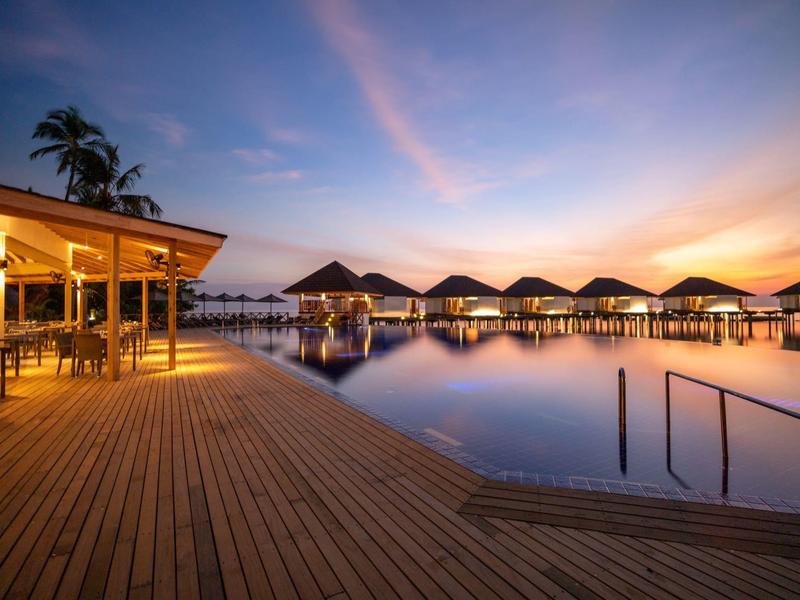 Evening view of overwater bungalows and a wooden deck with pool lighting.
