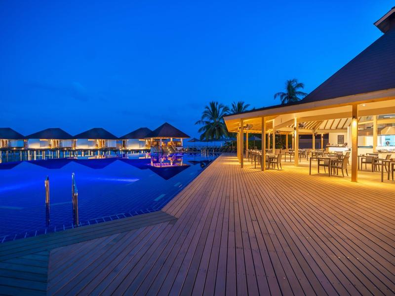 Evening view of a lit poolside restaurant with wooden deck and overwater bungalows.