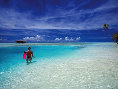 Osoba v čisté mělčině mořské vody pod modrou oblohou s několika mraky a palmami na okraji.