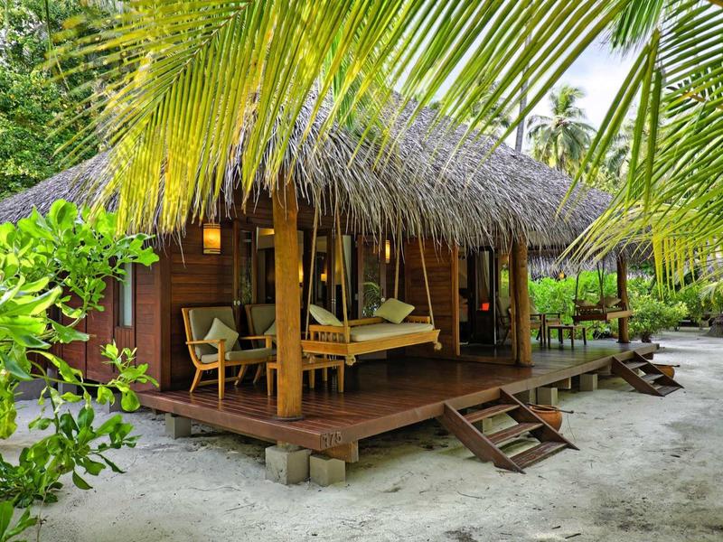 Dřevěný bungalov se střechou z došek na písečné pláži, obklopený palmami a tropickou vegetací.