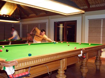 Zwei Männer spielen Billard in einem Raum mit Holzdecke und Fensterläden.