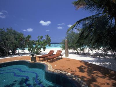 Area piscina con due lettini, circondata da palme e spiaggia di sabbia bianca sotto cielo azzurro.