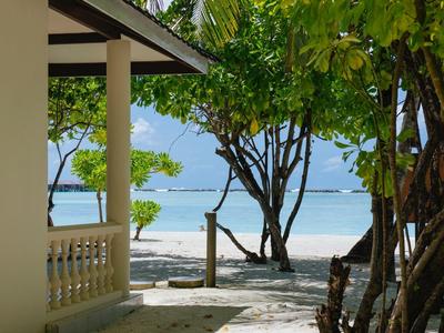 Blick vom Schatten eines Hauses zum Sandstrand mit Palmen und blauem Meer im Hintergrund.