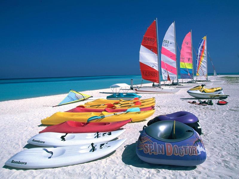 Bunte Surfbretter und Katamarane stehen auf weißem Sandstrand vor türkisfarbenem Meer und blauem Himmel.