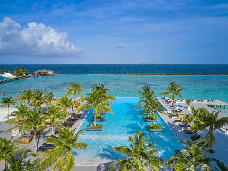 Tropischer Hotelpool mit Palmen, blauer Lagune und klarem Himmel im Hintergrund.