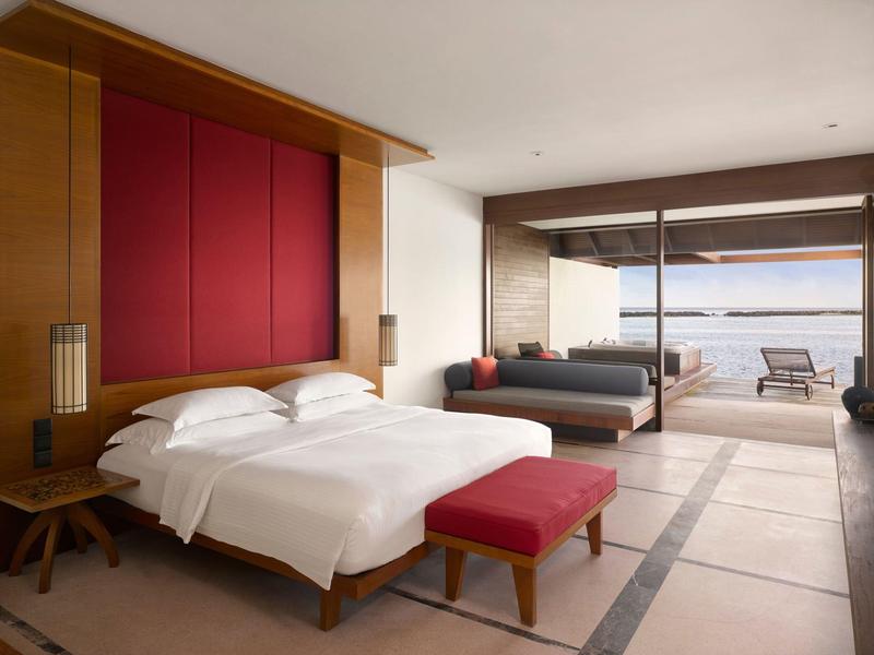 Modernes Schlafzimmer mit großem Bett, rotem Polster, Holzvertäfelung und Blick auf Terrasse mit Meer.