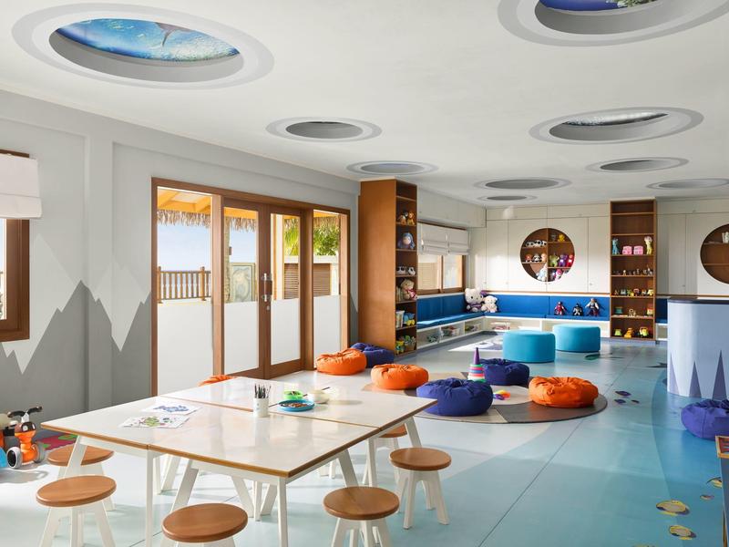 Hell eingerichteter Raum mit bunten Sitzkissen, Holzstühlen, großen Fenstern und runden Deckenfenstern.