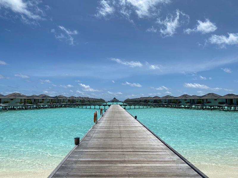 Langer Holzsteg führt zu über Wasser gebauten Bungalows in klarem, türkisfarbenem Meer unter blauem Himmel.