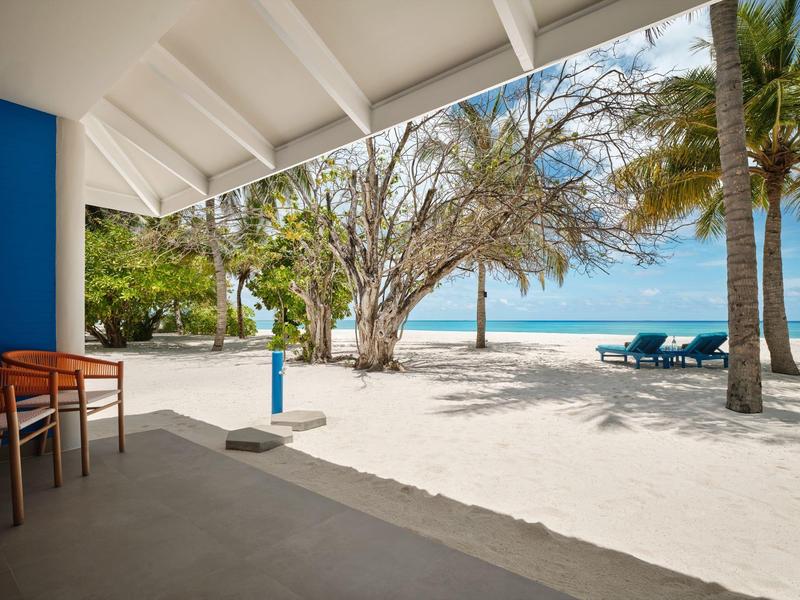 Strand mit weißem Sand, Palmen, blauer See und Terrasse mit Stühlen unter weißem Dach.