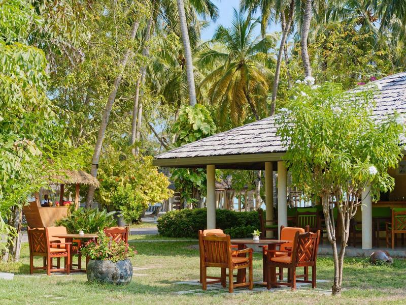 Gemütliche Sitzgelegenheiten mit Holztischen und Stühlen auf einer Rasenfläche neben einem Pavillon in tropischer Umgebung.
