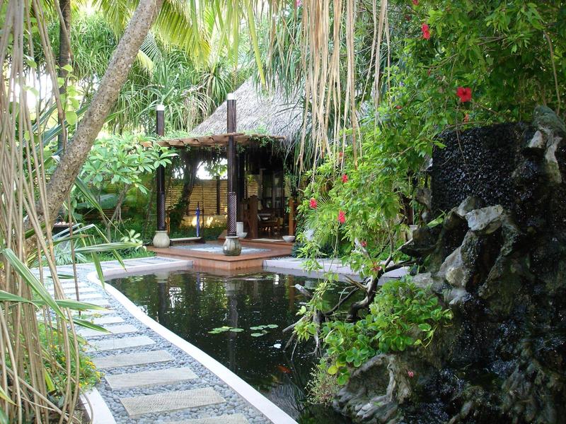 Ein tropischer Poolbereich mit Wasserfall, umgeben von üppiger grüner Vegetation und einem Holzpavillon.