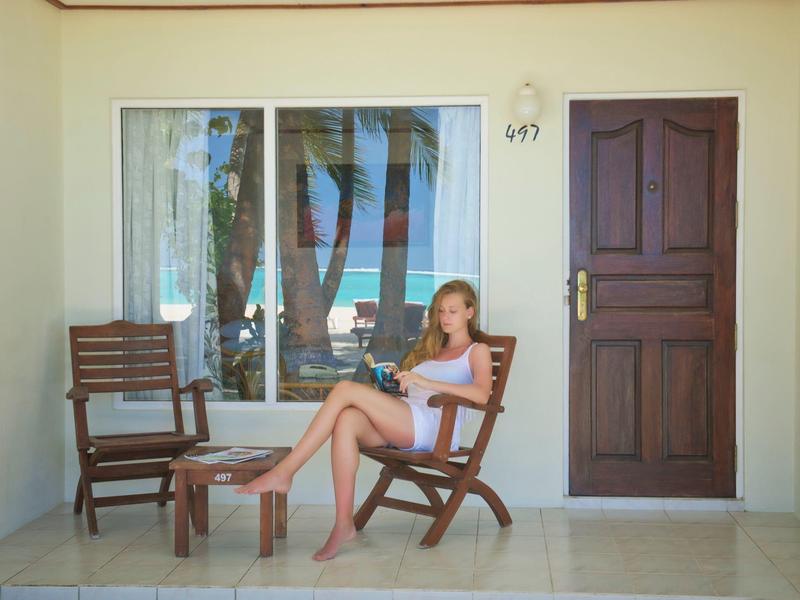Entspannte Frau sitzt auf Holzstuhl vor Hotelzimmer mit Palme im Hintergrund.
