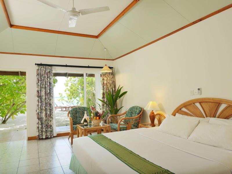 Helles Hotelzimmer mit großem Bett, grüner Pflanze und Blick auf die Terrasse mit Bäumen.