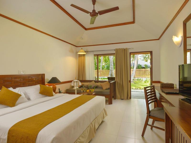 Helles Hotelzimmer mit Doppelbett, Holzstuhl, Fernseher und Blick auf Garten durch große Fenster.