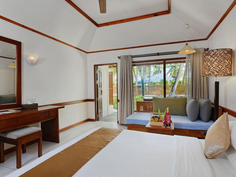 Helles Hotelzimmer mit großem Bett, Sitzbereich und Blick auf den Garten durch offene Türen.