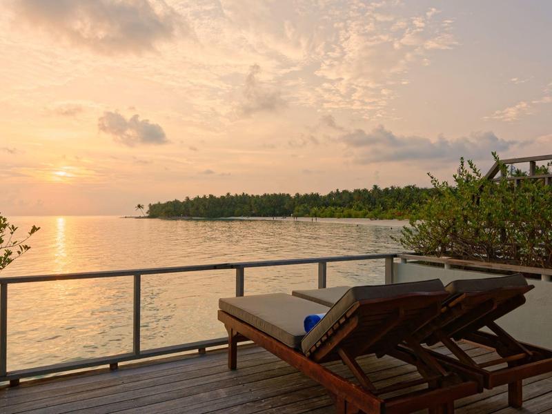 Zwei Liegestühle auf einem Balkon mit Blick auf einen Sonnenuntergang über dem Meer und einer bewaldeten Küste.
