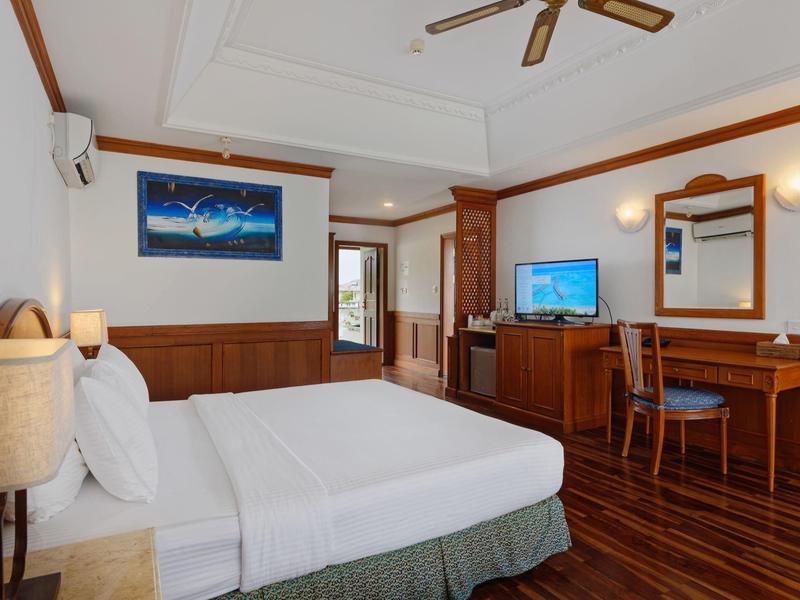 Helles Hotelzimmer mit Doppelbett, Holzmöbeln, Deckenventilator und Fernseher.