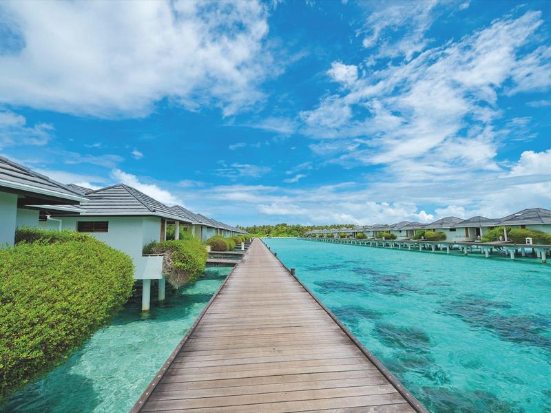 Holzsteg führt durch türkisfarbenes Wasser zu Überwasser-Bungalows unter blauem Himmel.