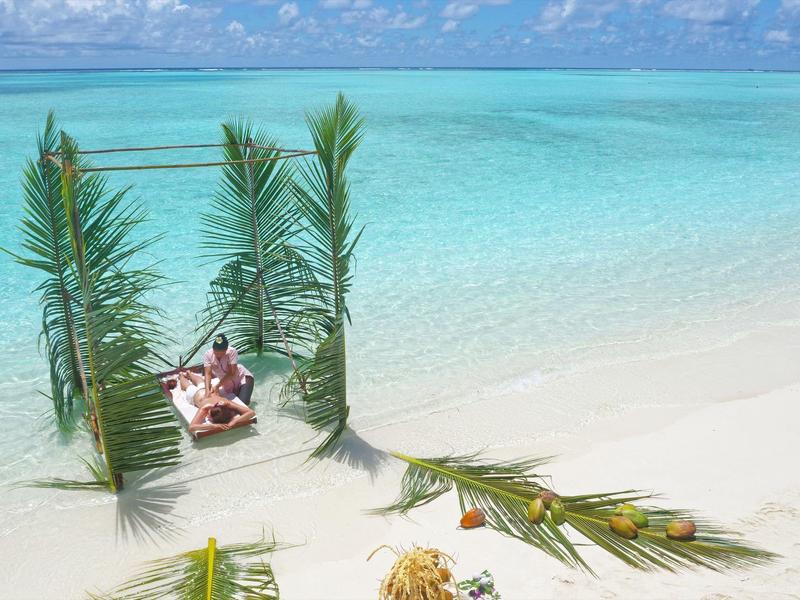 Strand mit türkisblauem Wasser, weißen Sand und Liegestühle unter Palmenzweigen.