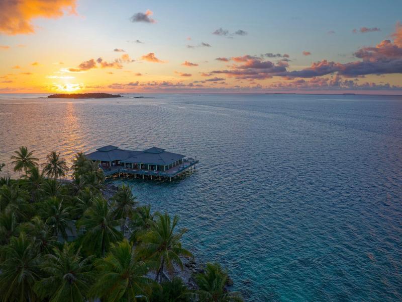 Sonnenuntergang über dem Meer mit Palmen und einem Haus auf Stelzen im Wasser.