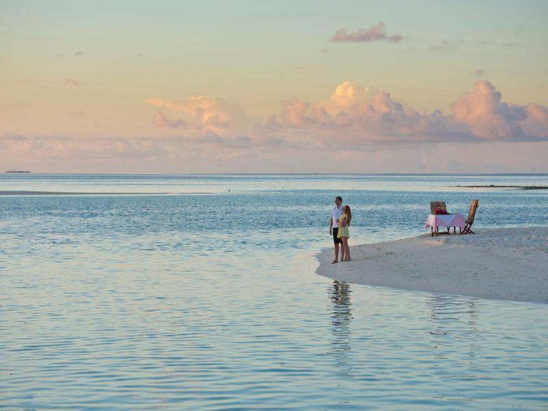 Person steht am Rand eines Sandstrands mit ruhigem Wasser und pastellfarbenem Himmel.