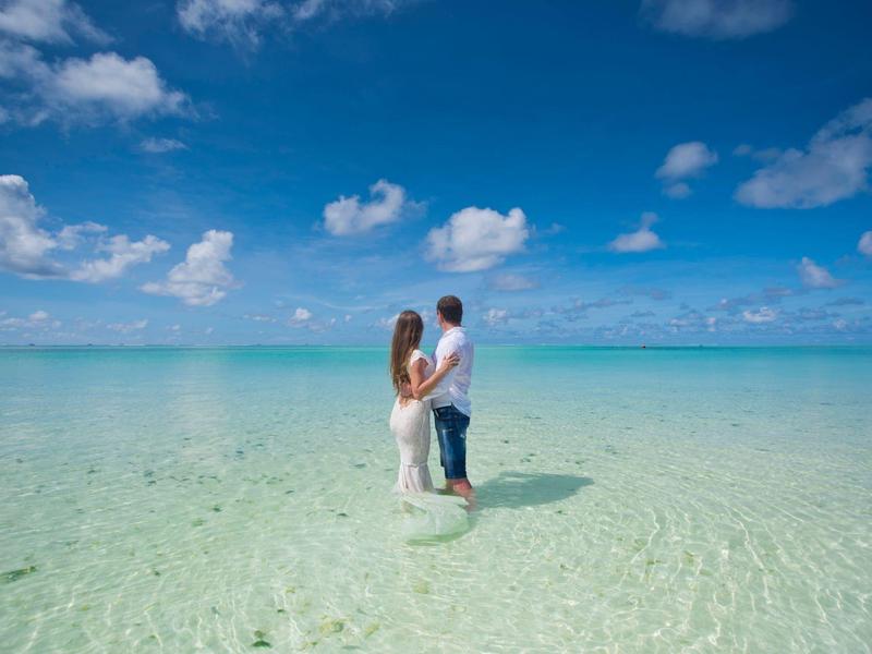 Paar steht im klaren, flachen Wasser eines tropischen Strandes unter blauem Himmel.