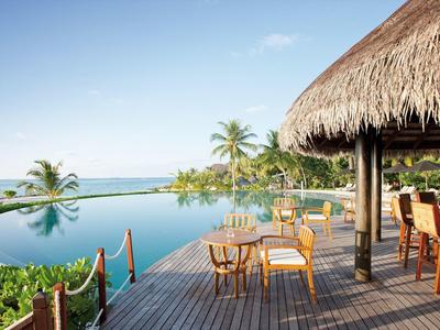 Infinity-Pool mit Holzdeck, Strohdeck und Sitzbereich, umgeben von Palmen und Meer im Hintergrund.