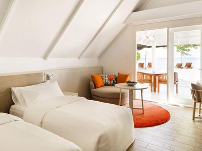 Helles Schlafzimmer mit zwei Einzelbetten, weißer Bettwäsche, orangem Teppich und gemütlicher Sitzecke.