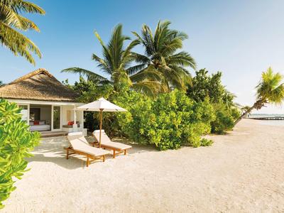 Strandvilla mit Liegen und Sonnenschirm, umgeben von Palmen und grünen Sträuchern bei klarem Himmel.