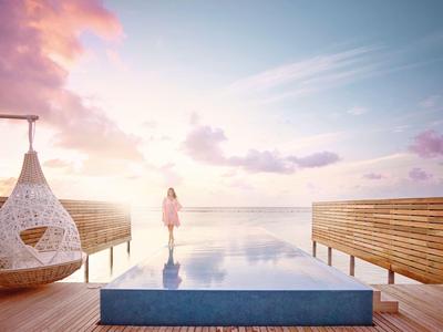 Une personne se tient au bout d'une piscine à débordement avec vue sur la mer au coucher du soleil.